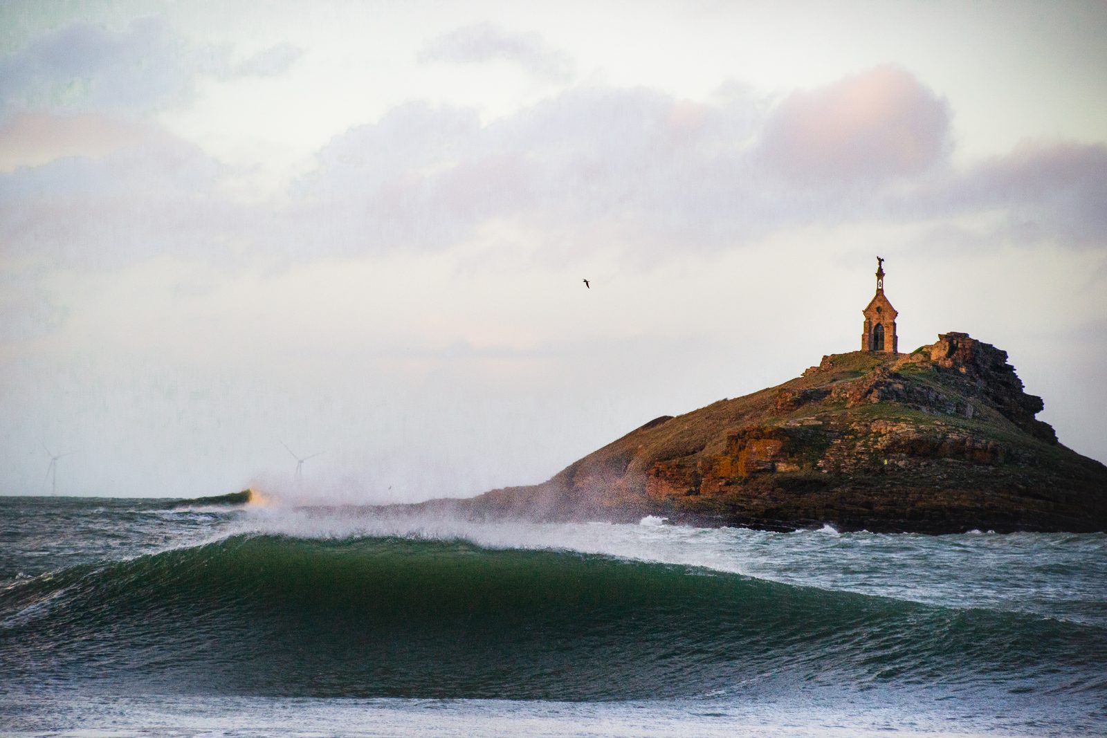 Une vague qui déroule devant un îlot rocheux surmonté d’une chapelle, sur la Côte d’Émeraude